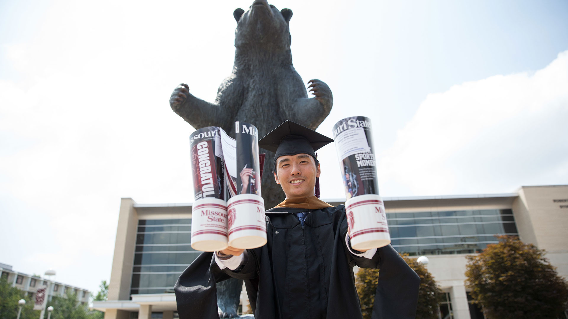 Missouri State graduate in cap and gown, holding up swag in front of bear statue. 